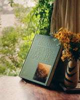 Fridays From The Garden cookbook props upright on a wooden table, framed by yellow wildflowers in a striped vase and trailing greenery beyond the window.