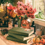Three copies of Fridays From The Garden stacked on a marble counter beside heirloom tomatoes, garlic, and a vase of pink poppies and snapdragons.