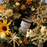 Flamingo Estate Golden Pollinator soy wax candle surrounded by a lush arrangement of yellow sunflowers and white blooms.