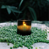 A lit Flamingo Estate Green Goddess Candle glows on a silver platter blanketed in bright green peas, with leafy foliage shadowed in the background — basil, mint and pea tendrils embodied.