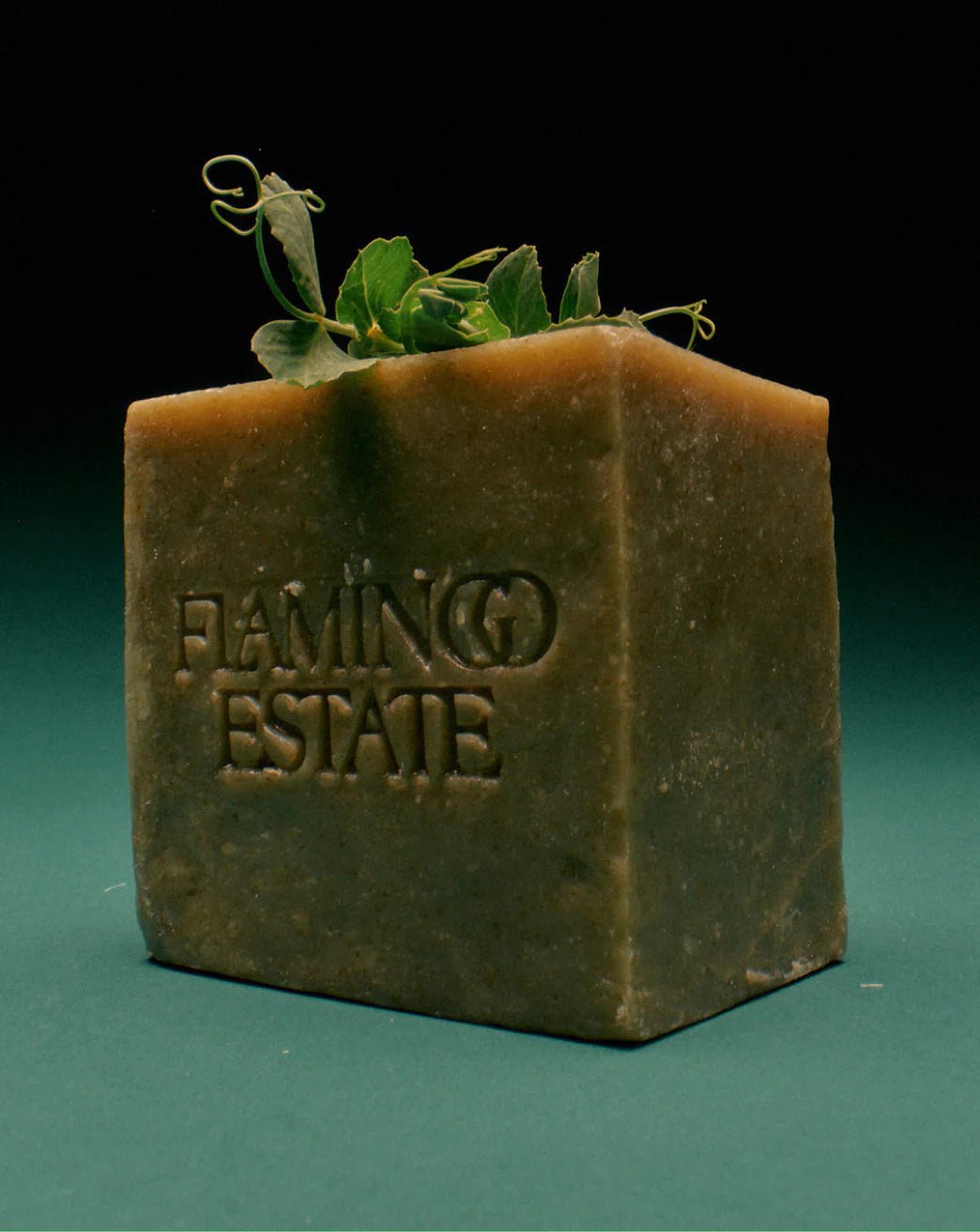 The tall olive-green Flamingo Estate Green Goddess Soap Brick stands upright with curling pea tendrils sprouting from its top against a deep emerald background.