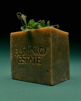 The tall olive-green Flamingo Estate Green Goddess Soap Brick stands upright with curling pea tendrils sprouting from its top against a deep emerald background.