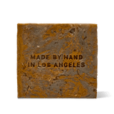 The reverse face of the Flamingo Estate Green Goddess Soap Brick, marbled in olive and bronze swirls, embossed with Made by Hand in Los Angeles on a soft grey backdrop.