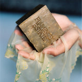 Pale hands wrapped in delicate cream lingerie embroidered with yellow flowers cradle the Flamingo Estate Green Goddess Soap Brick against a soft blue backdrop.