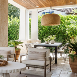 Outdoor patio furniture set with woven wicker chairs, beige cushioned armchairs, a round white table, and a dining table under a covered patio with stone columns and greenery.