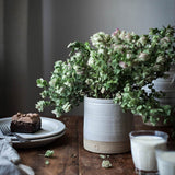 Farmhouse Pottery  Silo Crock used as a vase with green foliage on a rustic wooden table next to a slice of chocolate cake.