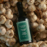 The Flamingo Estate Heirloom Garlic olive oil bottle lies across a bed of whole garlic bulbs and papery skins in moody, low light.