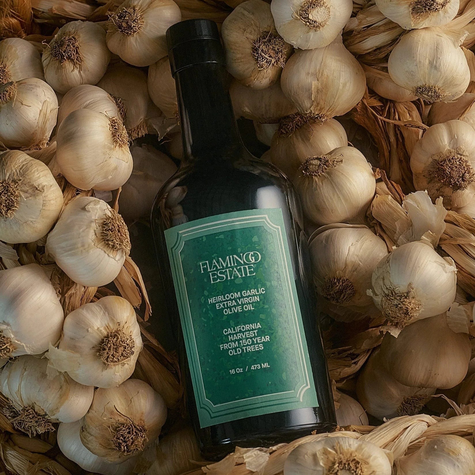 The Flamingo Estate Heirloom Garlic olive oil bottle lies across a bed of whole garlic bulbs and papery skins in moody, low light.