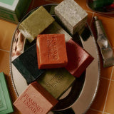 An assortment of Flamingo Estate soap bricks in red, black, olive and grey-speckled tones piled on a silver tray atop terracotta tile, with a hand cream tube alongside.