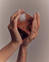 Manicured hands lather the rust-red Flamingo Estate Heirloom Tomato Soap Bar between sudsy palms against a warm beige background.