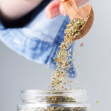 Calicutts Herbes De Provence spice blend being poured from a wooden spoon into a glass jar.