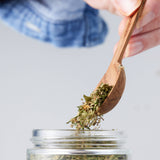 Calicutts Herbs Blend being poured from a wooden spoon into a glass jar, showcasing dried green aromatic herbs.
