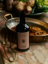 Heritage Extra Virgin Olive Oil rests on a marble counter beside a copper pan of roasted vegetables and fresh produce in a candlelit kitchen.