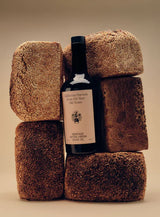 A Flamingo Estate Heritage olive oil bottle nestles between sculptural loaves of seeded brown bread stacked against a warm sand-toned backdrop.