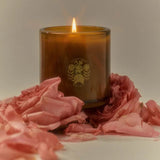 A brown glass Night Blooming Jasmine & Damask Rose Candle from Calimia Home Shop sits amid pink rose petals, its flame aglow. The front shows a subtle flamingo and foliage logo, set against a softly lit, neutral background.