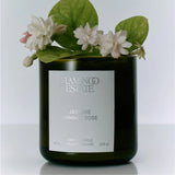 A green glass candle jar labeled FLAMINGO ESTATE Night Blooming Jasmine & Damask Rose, evoking the Euphoria fragrance, topped with white flowers and green leaves, is set against a light neutral background.