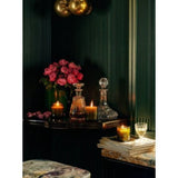 A cozy corner with a Night Blooming Jasmine & Damask Rose Candle glowing, a rose bouquet in a vase, crystal decanters, white wine, and stacked books on a marble table, framed by dark paneled walls and brass light fixtures.