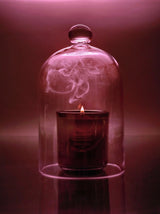 A Night Blooming Jasmine & Damask Rose Candle glows beneath a glass dome with swirling smoke, bathed in soft pink light for a warm, atmospheric scene.