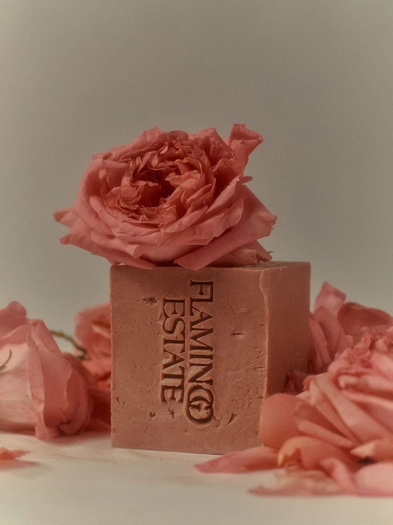 A pink Flamingo Estate Jasmine Damask Rose Soap Brick stands upright crowned with a fluffy blooming pink rose, surrounded by other dusty rose petals.