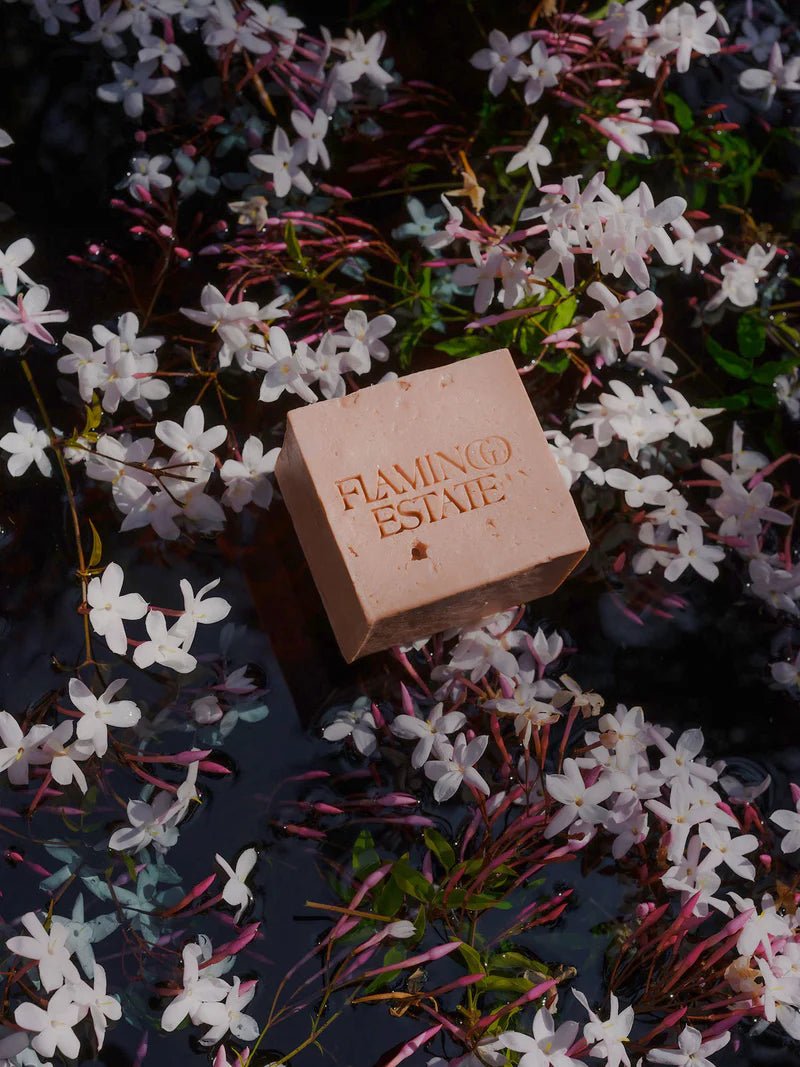 The pink Flamingo Estate Jasmine Damask Rose Soap Brick floats among delicate white jasmine blossoms scattered across dark moody pond water.