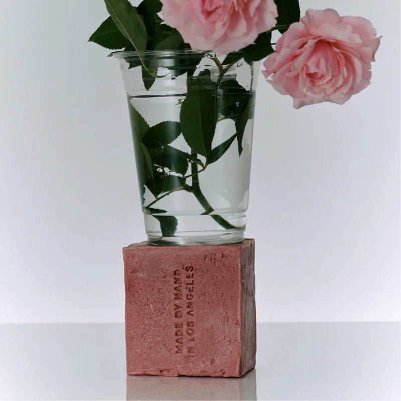 A clear glass tumbler of pink garden roses sits balanced atop the rose-pink Flamingo Estate Jasmine Damask Rose Soap Brick embossed Made by Hand in Los Angeles.