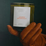 Flamingo Estate Leather Appointment scented candle in a forest-green fluted glass held by a hand in a tan leather glove.