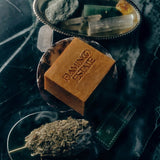 Flamingo Estate Manuka honey soap brick with embossed logo on a dark marble surface with dried herbs and crystals.