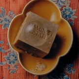 Flamingo Estate Manuka honey soap brick in a yellow ceramic dish on a floral patterned fabric surface.