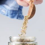 Calicutts Mediterranean spice blend being poured from a wooden spoon into a glass jar with visible dried herbs
