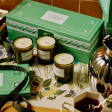 The Mini Three Sisters Candle Gift Set styled on terracotta tile beside a silver teapot and floral cup, three candles arranged in front of the green Flamingo Estate box.