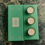 Top-down view of the open green Flamingo Estate gift box revealing three creamy candle inserts beside the closed lid on a moody green marble surface.
