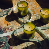 Three lit Flamingo Estate Three Sisters candles flicker in green glass jars on a marbled surface, draped with the brand's signature ivory ribbon.