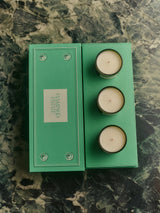 Top-down view of the open green Flamingo Estate gift box revealing three creamy candle inserts beside the closed lid on a moody green marble surface.