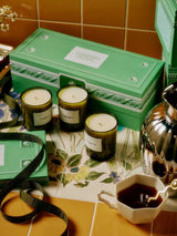The Mini Three Sisters Candle Gift Set styled on terracotta tile beside a silver teapot and floral cup, three candles arranged in front of the green Flamingo Estate box.
