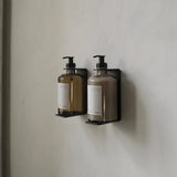Calimia Home matte black wall-mounted bottle holder brackets holding two pump bottles on a neutral wall.