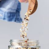 Calicutts Montreal spice blend being poured from a wooden spoon into a glass jar with visible peppercorns and salt.