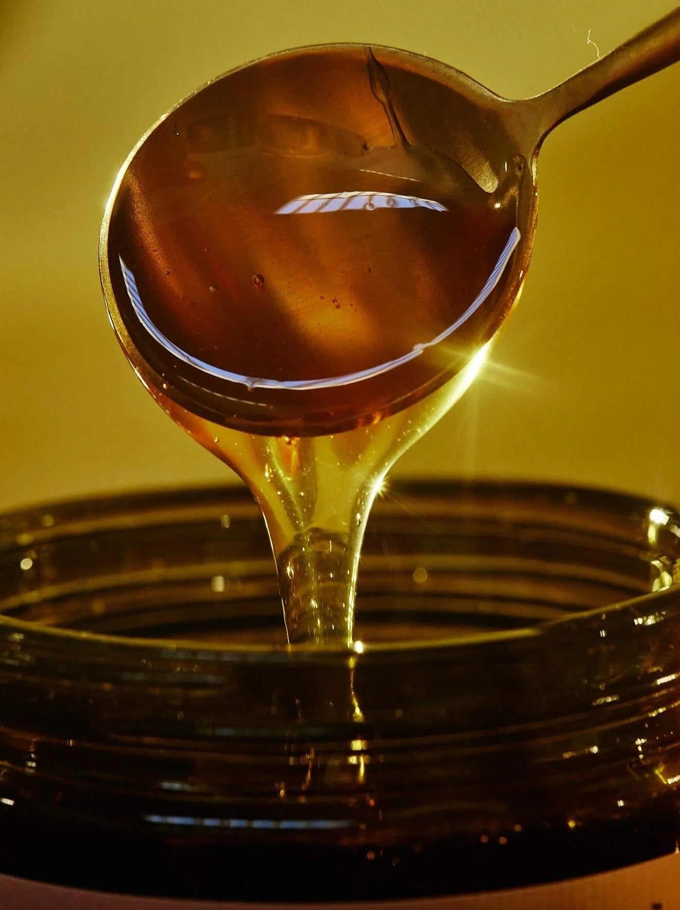 A brass ladle pours a glistening stream of Flamingo Estate Morita Chile honey into a wide jar in a richly backlit, golden-hued shot.