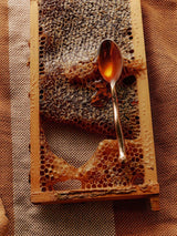 A wooden bee-frame holds raw honeycomb on jute, a brass spoon catching golden Flamingo Estate California wildflower honey.