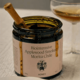 An open Flamingo Estate Applewood Smoked Morita Chile Honey jar with a wooden dipper inside, a coupe of amber wine alongside on linen.