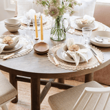 Valencia X Dining Table by Grove & Anchor in natural oak, round top styled with a full place setting of woven placemats, linen napkins in straw rings, plates and candles