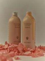 Cream Flamingo Estate Euphoria Hand Soap and Body Wash bottles stand on a bed of pink petals, lit with soft warm light for a romantic mood.