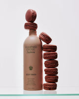 A taupe Flamingo Estate Euphoria Body Wash bottle balances a stack of dark mauve macarons against a clean white backdrop, playful and sweet.