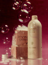 A cream Flamingo Estate Euphoria Body Wash bottle stands beside a foamy stamped soap bar amid floating bubbles on a rich burgundy background.