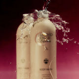 Two cream Flamingo Estate Euphoria Body Wash bottles are caught mid-splash with a burst of water against a deep magenta backdrop.