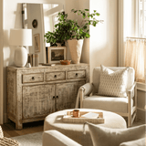 A styled living room interior featuring a large woven dome pendant light hanging over a distressed wood credenza, with a cream armchair, round ottoman, and potted green plants in a bright, sunlit room.