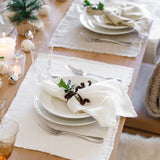 Pom Pom At Home ivory linen-cotton placemats with frayed edges set on a wooden dining table with white plates and napkins.