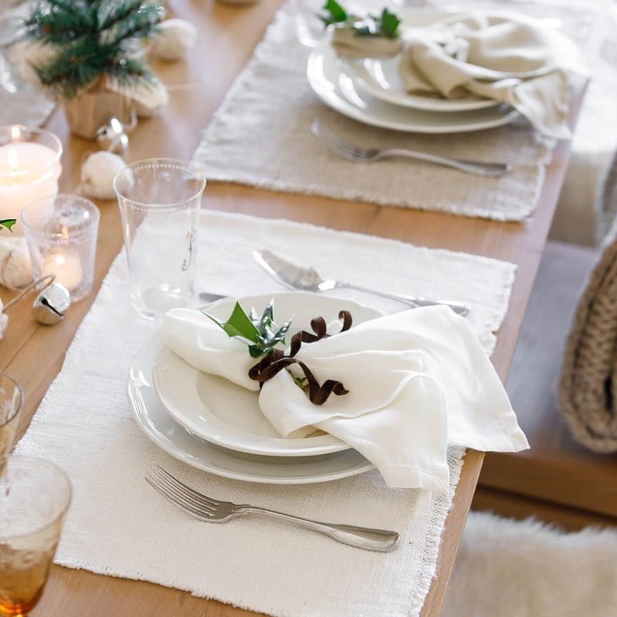 Pom Pom At Home ivory linen-cotton placemats with frayed edges set on a wooden dining table with white plates and napkins.