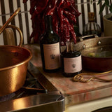 The Tablemates Heritage Extra Virgin Olive Oil and Balsamic Vinegar pose on a marble counter beside a copper pot, with a cluster of dried red chiles hanging above.