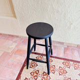 Calimia Home Omah Stool in black stained wood with a round seat and tapered legs on a tiled floor