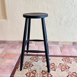 Calimia Home Omah stool in black stained wood with a round seat and tapered legs on a patterned tile floor.